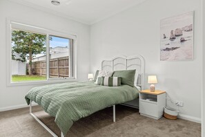 Ein Schlafzimmer mit Bett, Nachttisch, Lampe und einem Fenster mit Blick auf einen Garten.