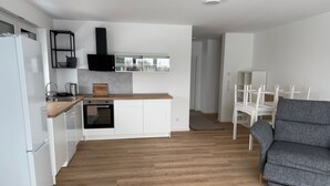 Et moderne køkken med hvide skabe, en træbordplade og en indbygget ovn. Der er en grå lænestol og hvide stole i det tilstødende opholdsområde.