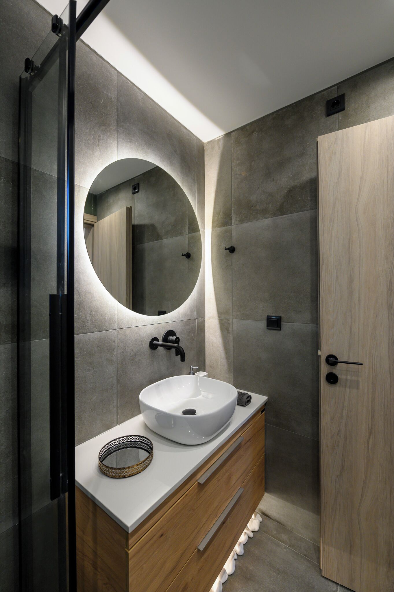 Een moderne badkamer met een ronde spiegel, een wit wastafelblad, een houten kastje en een douchecabine met glazen wanden.