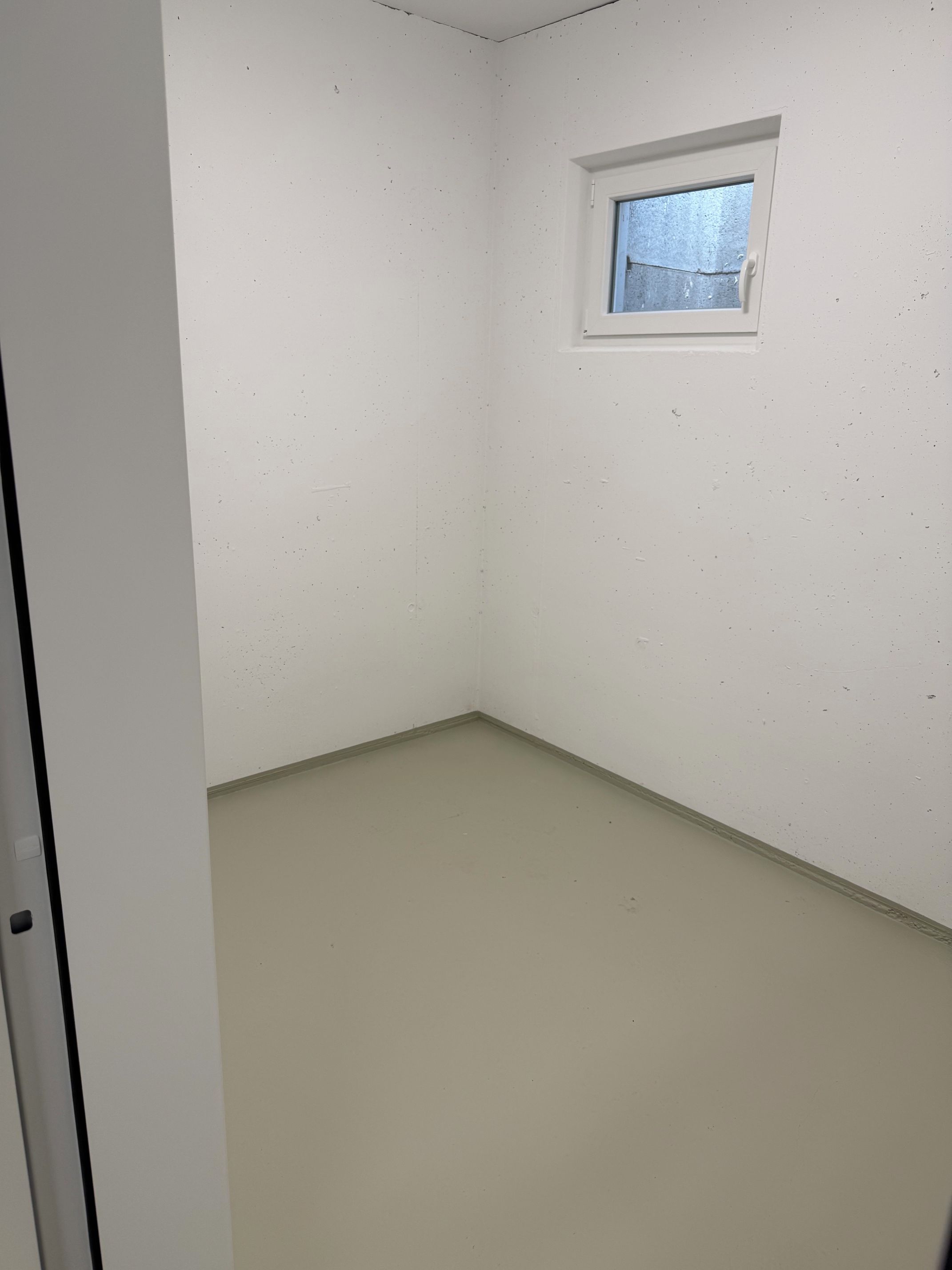 Ein Zimmer mit einem kleinen Fenster, weißen Wänden und einem schlichten Boden.