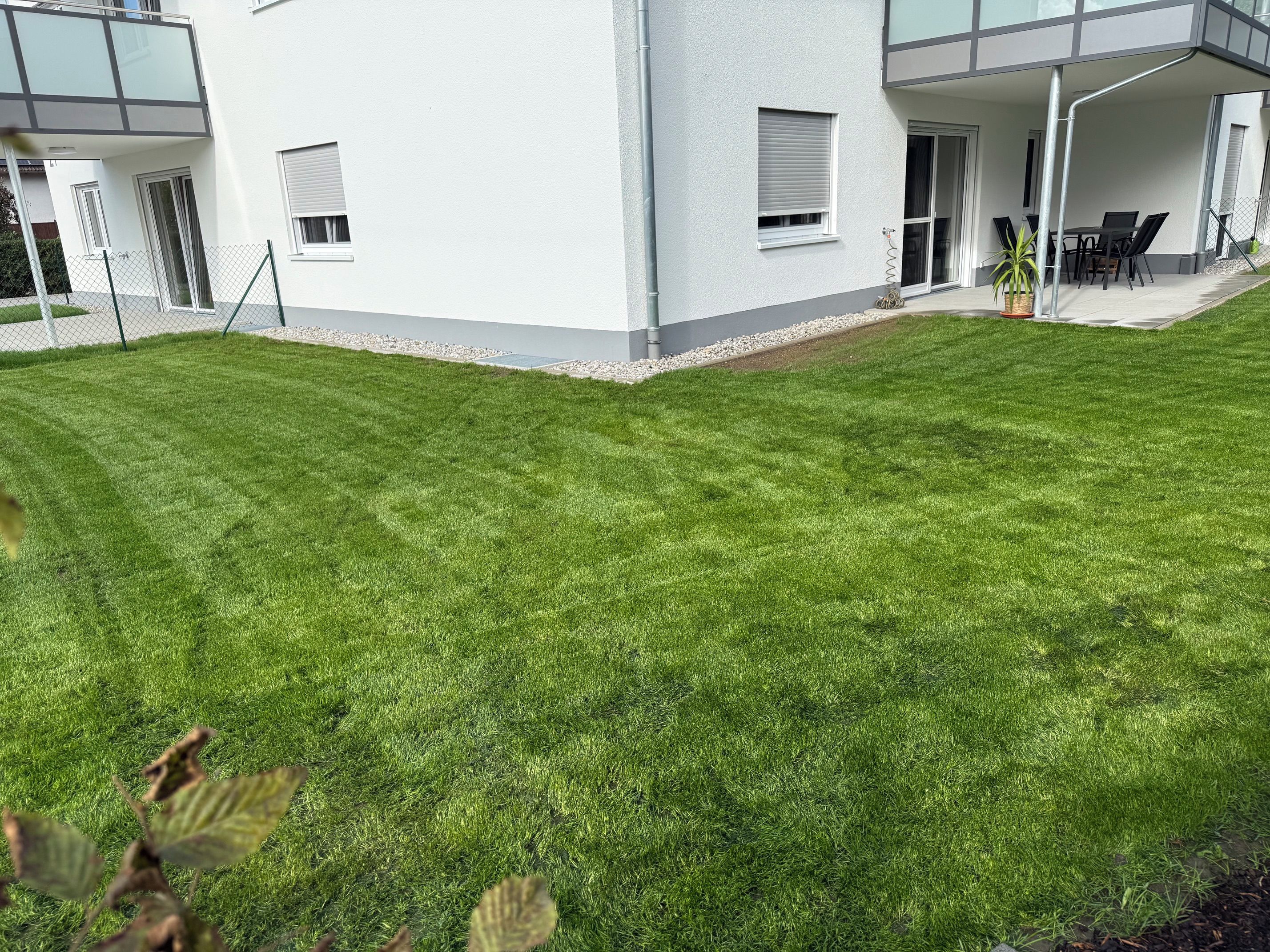 Ein modernes Gebäude mit gepflegtem Rasen, einer Terrasse und Gartenmöbeln, sowie einem wolkenlosen Himmel.