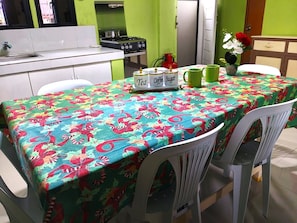 A dining area with a table covered in a floral tablecloth, chairs, a kitchen in the background, and a vase with flowers.