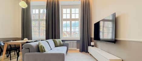Una sala de estar moderna con sofá, televisión, mesa de comedor y ventanas grandes con cortinas.