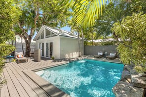 Ein Garten mit Schwimmbad, einer Holzterrasse und einem kleinen weißen Gebäude mit Glasüren.