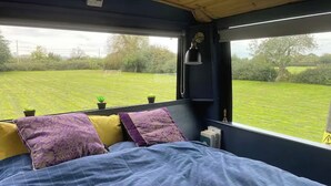Cama com colcha azul, duas almofadas roxas e vista para um campo verde através de uma janela.