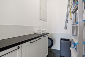Ein Badezimmer mit schwarzer Ablagefläche, weißen Schränken, einem Spiegel, einer Waschmaschine und einem Mülleimer.