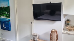 En stue med et fladskærms-tv ophængt på væggen, en dør, et lille trækonsolbord med bøger og pyngeting, samt et maleri af et bybillede.