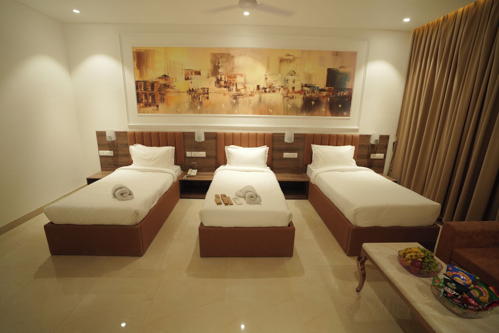 A hotel room with three single beds, a large abstract painting on the wall, and a dining table with snacks.