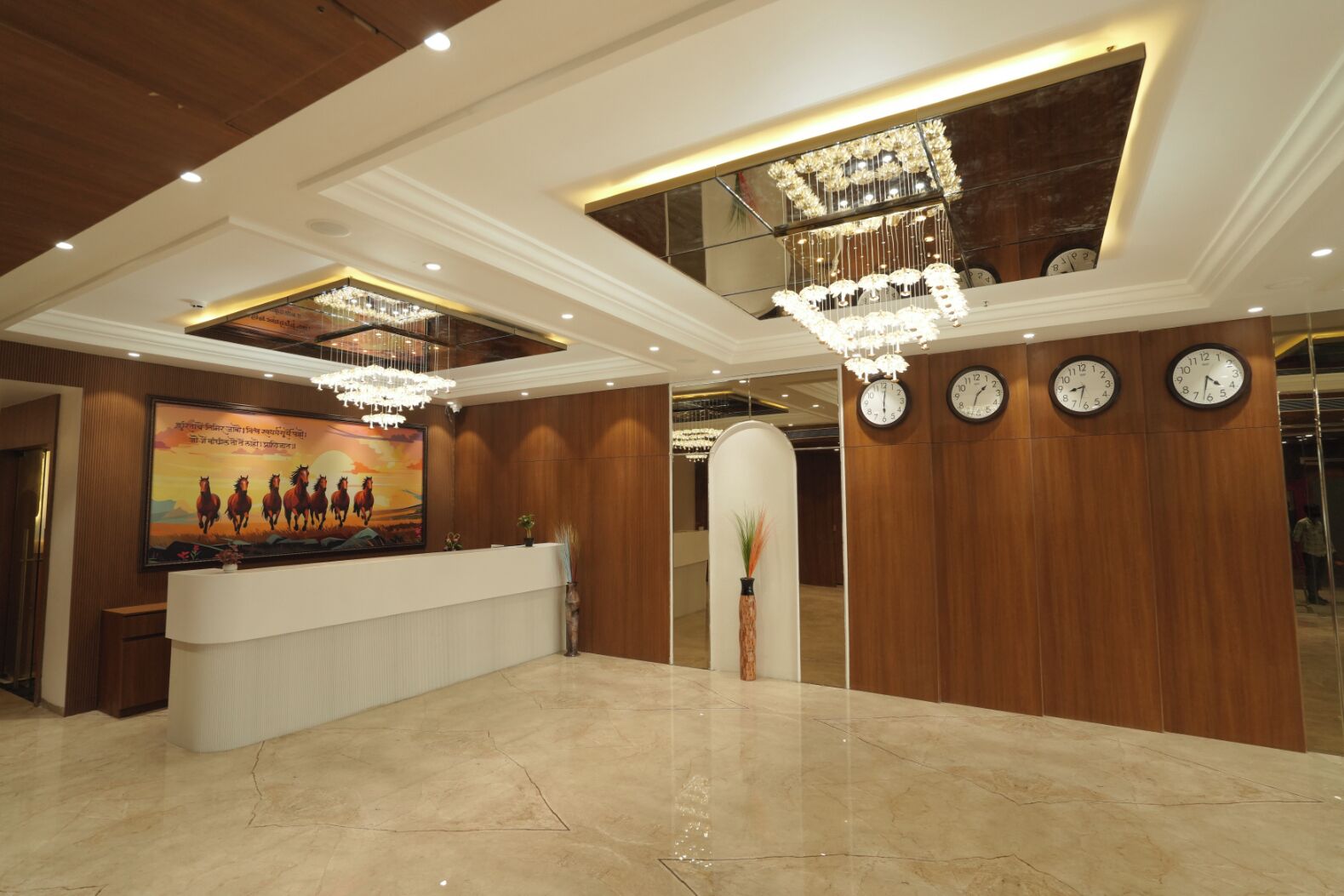 Un'area reception di un hotel con pavimento in marmo, pannelli in legno, una scrivania della reception, un grande poster, lampadari e orologi alle pareti.