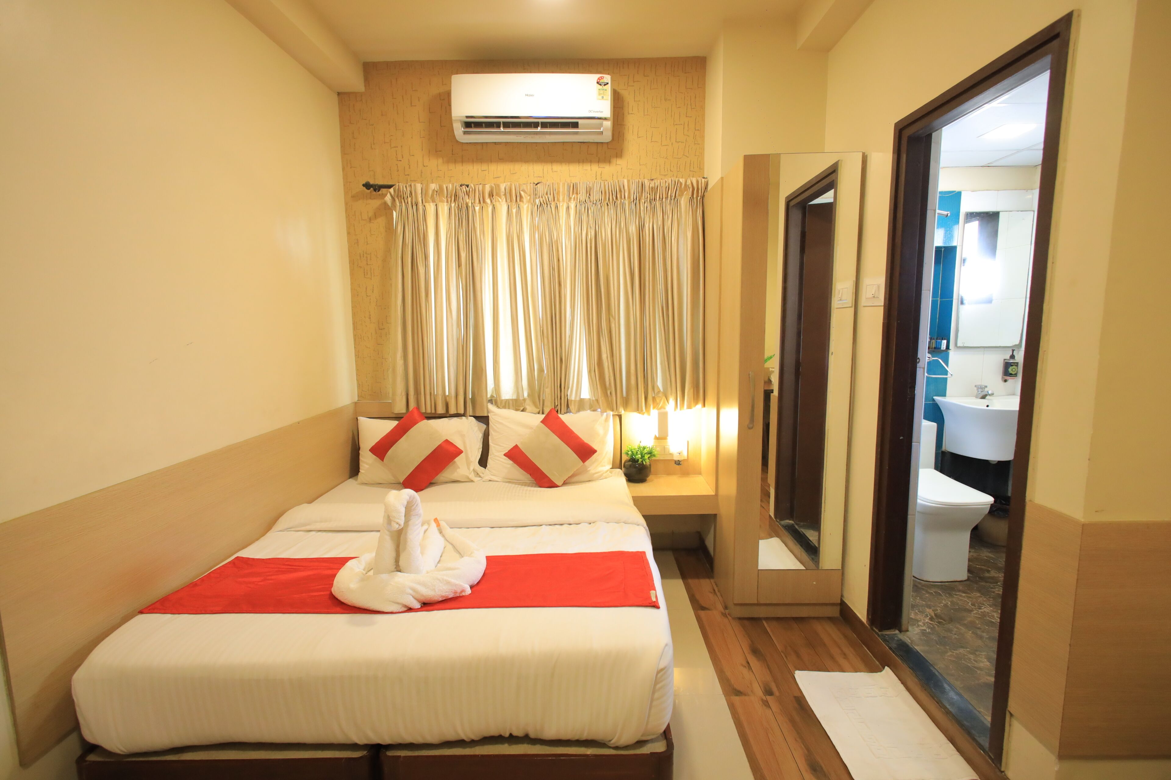 A hotel room with a bed, a bedside table with a lamp, a window with curtains, and a bathroom visible through an open door.