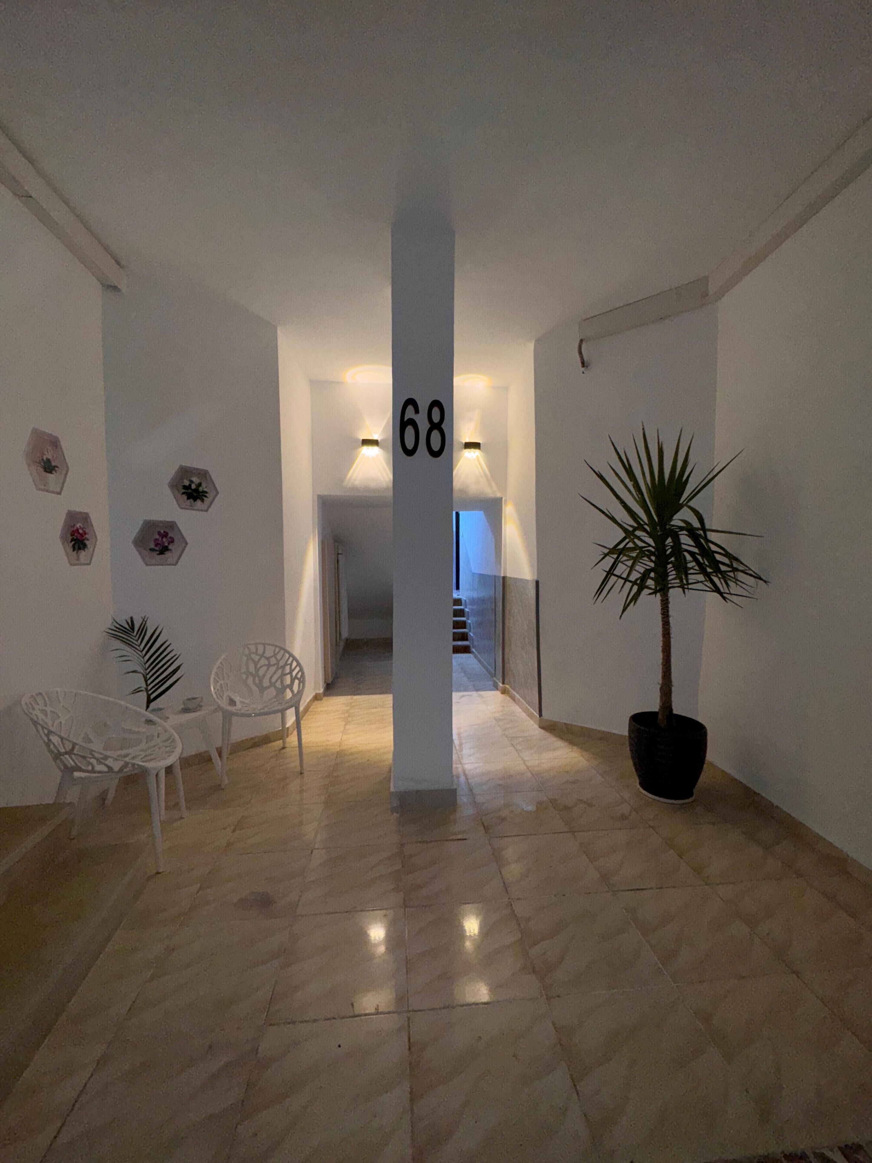 A hallway with a number sign, a potted plant, two chairs, and a staircase in the background.