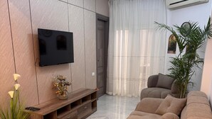 Una sala de estar moderna con un amplio sofá modular, un televisor de pantalla plana, un mueble de entretenimiento de madera, una planta en maceta y un gran ventanal con cortinas.