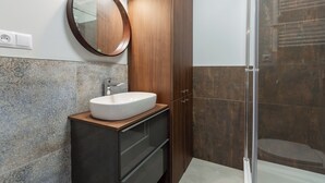 Een badkamer met een ronde spiegel, een wit wastafelmeubel, donkere kastjes en een douchehoek.
