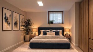 Quarto com uma cama grande, mesas de cabeceira, janela com cortinas, planta em vaso e obras de arte emolduradas na parede.