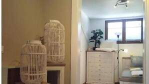 Um quarto com piso de madeira, uma cômoda branca, uma mesinha de cabeceira e duas gaiolas de pássaros brancas em suportes.