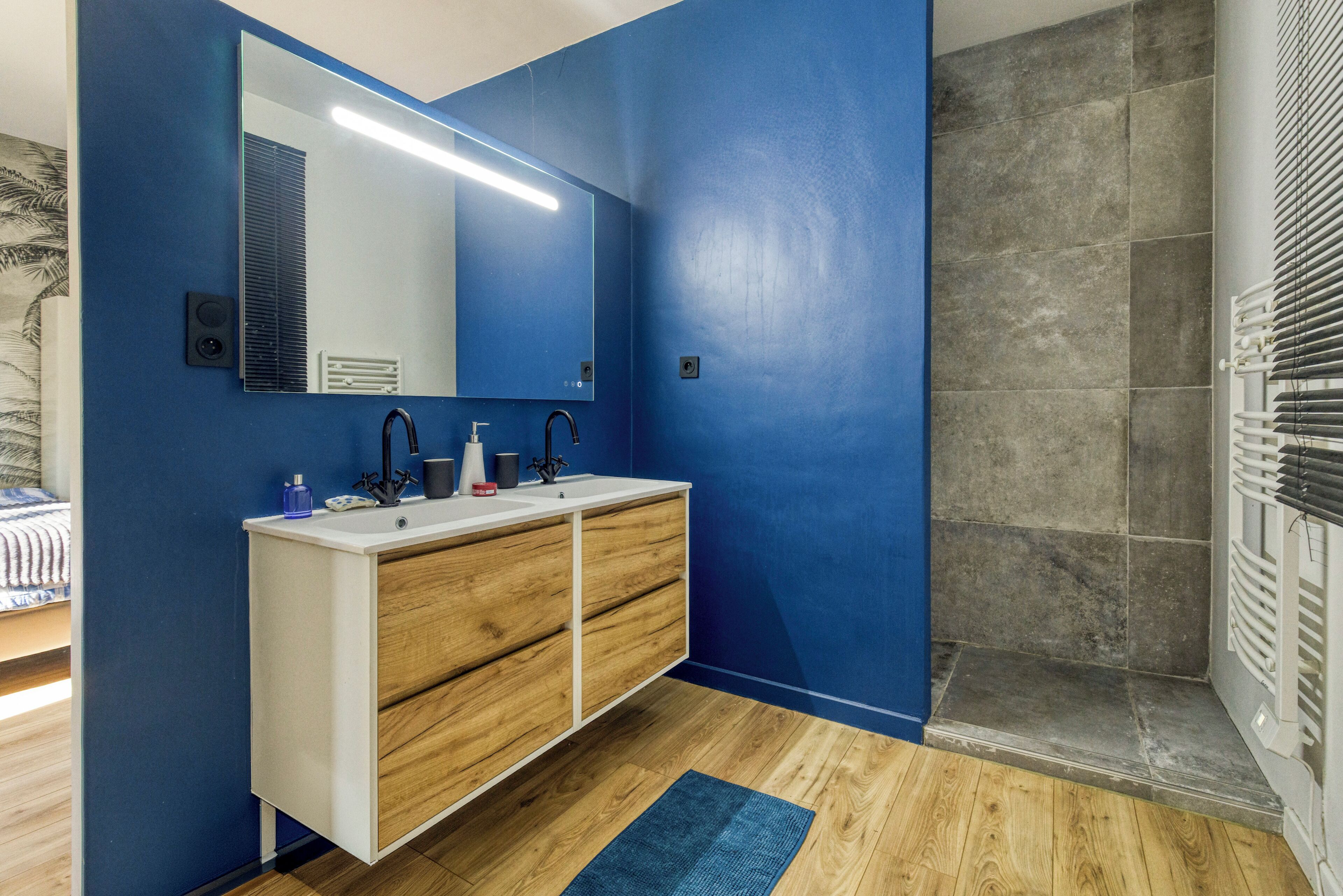 Un bagno con una parete blu, un lavandino bianco con cassetti in legno, uno specchio e uno spazio doccia.