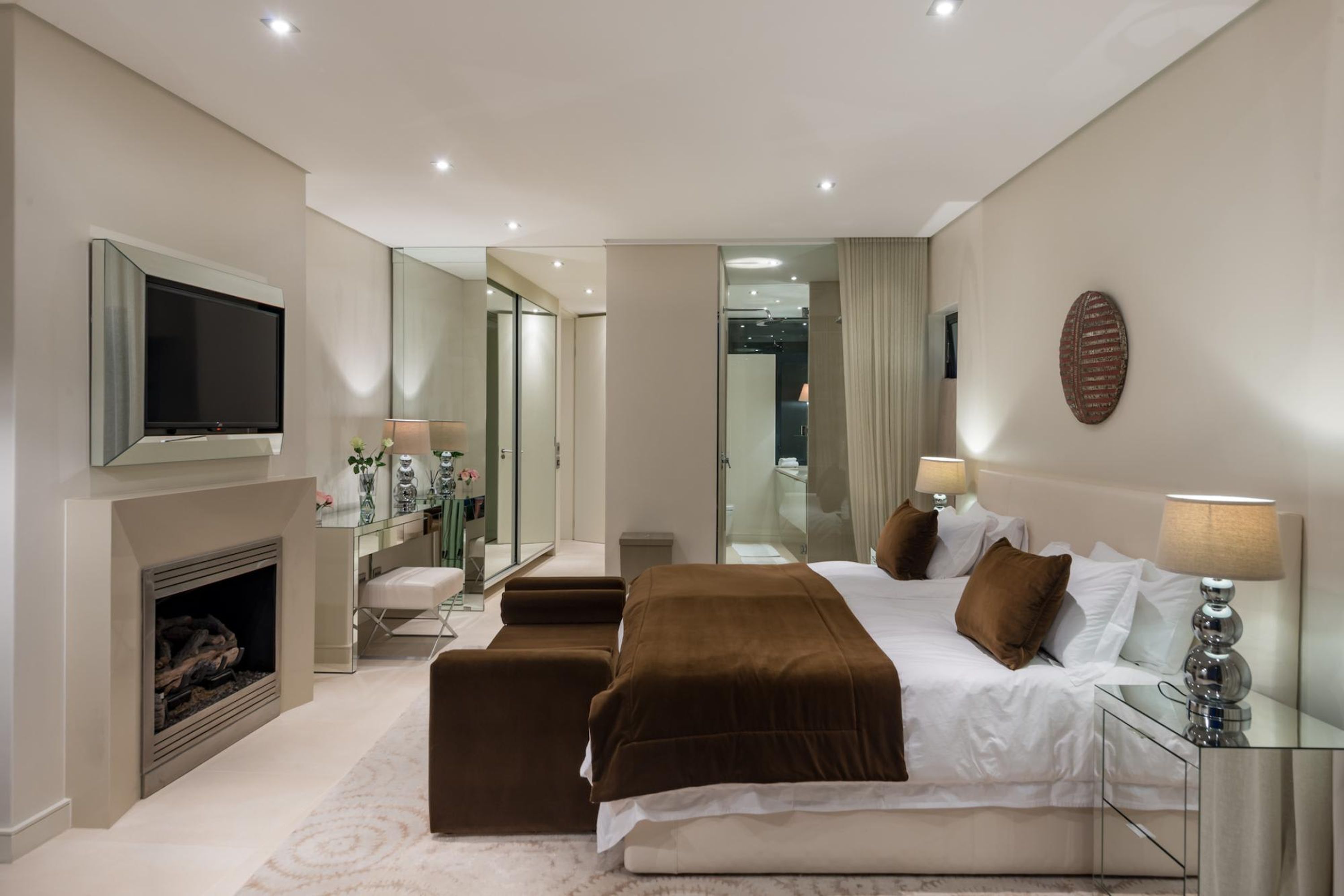 A modern hotel room with a large bed, a flat-screen TV, a fireplace, a desk, and a mirror.