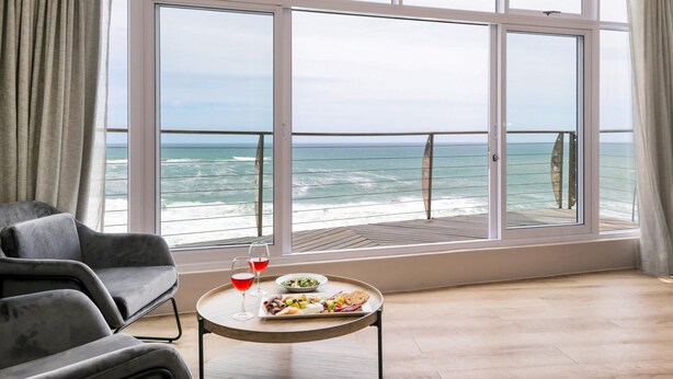 A room with a large window overlooking the ocean, a dining table with food and drink, and a grey sofa. - Camps Bay 6bed w Ocean Views and Pool Villa Sevara (Cape Town)
