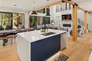 A modern kitchen with a large island, wooden beams, a fireplace, and a dining area with a view of the forest. - Memories Are Made Here | Chef's Kitchen & Prime Location (Cle Elum)