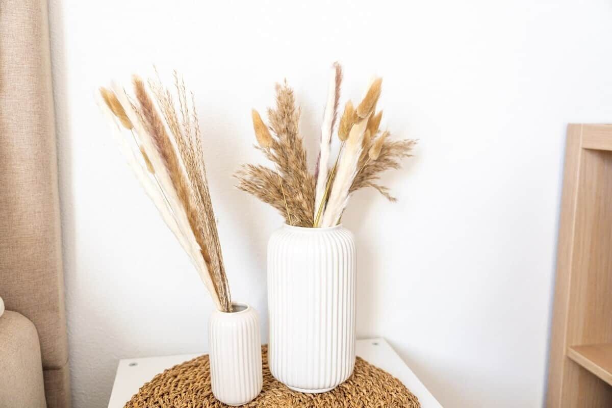 Two white vases with dried plants on a white table against a plain wall.