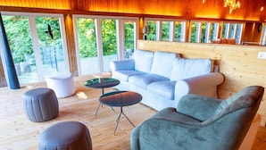 Quarto com sofá, pufes, uma mesa pequena e uma grande janela com vista para o verde.