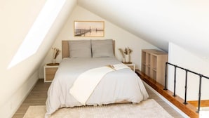 Quarto com teto inclinado, cama com edredom branco, mesa de cabeceira com um vaso e um quadro na parede.