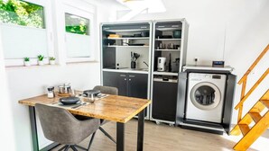 Uma cozinha compacta com uma mesa de madeira, uma cadeira cinza, uma máquina de lavar e um forno embutido.