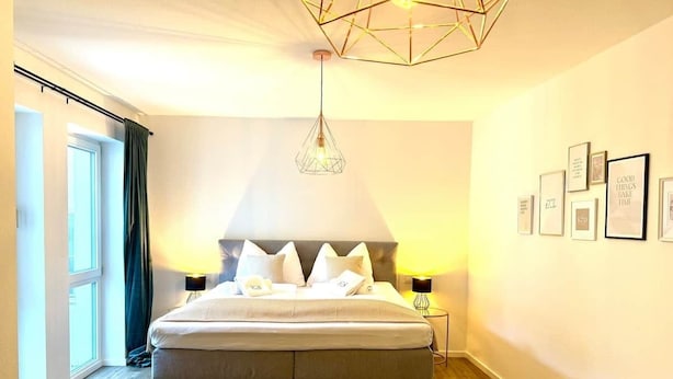 A modern bedroom with a large bed, two bedside lamps, a geometric ceiling light, and a wall with framed pictures. - VetterLux Bellevue, suite, citycenter Ravensburg (Ravensburg)