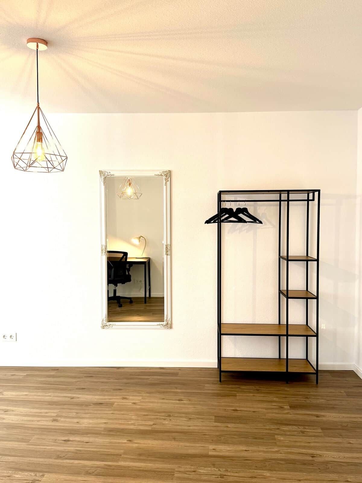 Una habitación con piso de madera, lámpara colgante, espejo y estantería metálica negra.