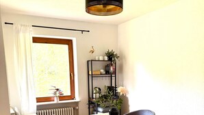 Un coin repas avec une table en bois, quatre chaises grises, une lampe noire suspendue, un radiateur, une fenêtre avec des rideaux blancs et une étagère avec des plantes et des ustensiles de cuisine.