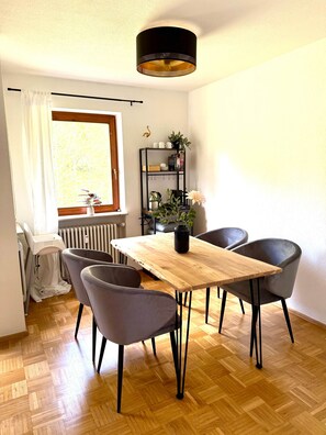 A dining area with a wooden table, four grey chairs, a black hanging lamp, a radiator, a window with white curtains, and a shelf with plants and kitchen items. - VetterLux Lumi - Balcony, Parking, Greenery view (Ravensburg)