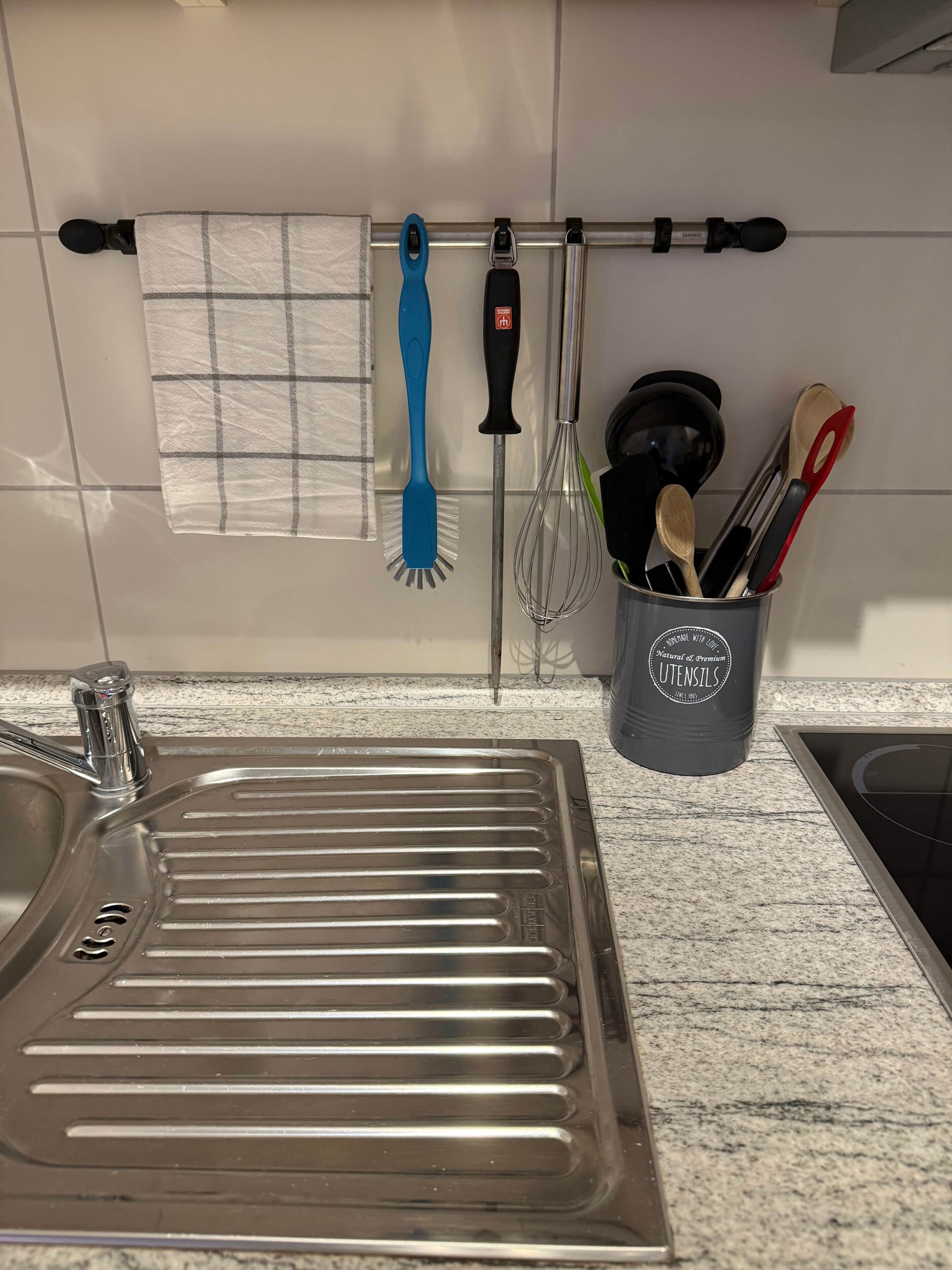A kitchen with a stainless steel sink, a dish rack, a hanging rack with cleaning tools, and a utensil holder.