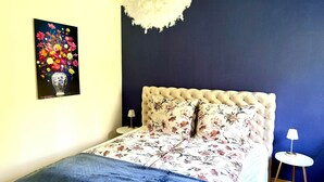Une chambre à coucher avec une tête de lit capitonnée, une literie à motifs floraux, une table de chevet avec une lampe et un tableau représentant des fleurs accroché au mur.