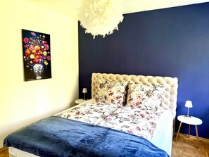 A bedroom with a tufted headboard, floral bedding, a side table with a lamp, and a painting of flowers on the wall. - VetterLux Lumi - Balcony, Parking, Greenery view (Ravensburg)