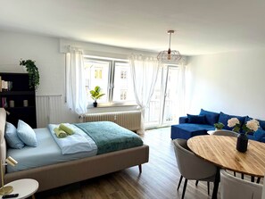 Een moderne hotelkamer met een bed, een ronde houten tafel, een blauwe bank en een raam met gordijnen.