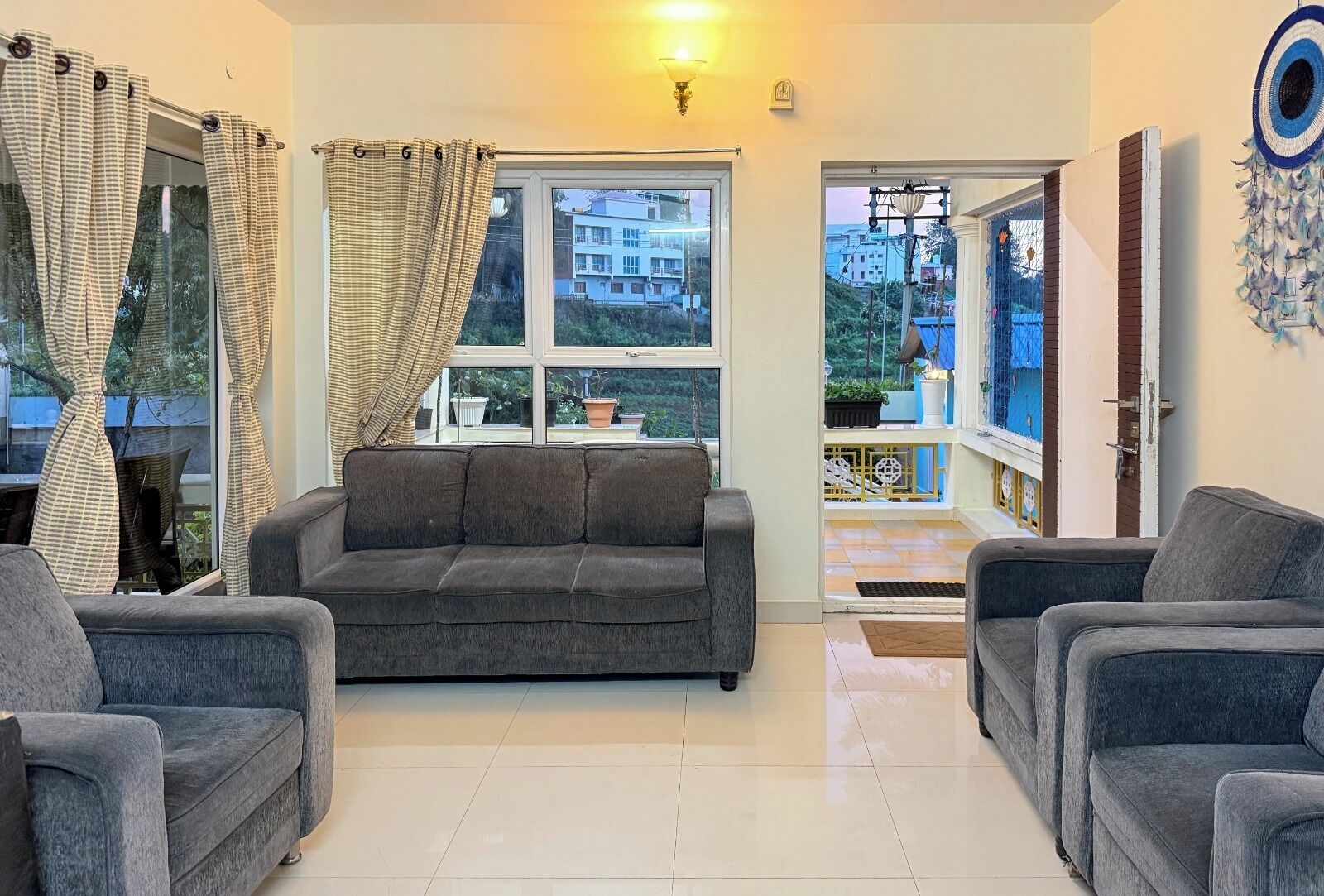 A living room with a grey sofa, two armchairs, a large window with curtains, and a view of buildings outside.