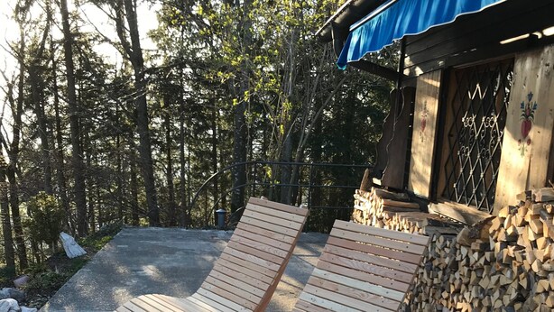 Deux fauteuils en bois sont disposés dans un espace extérieur ensoleillé, à côté d’une pile de bois de chauffage. On aperçoit en arrière-plan un bâtiment surmonté d’un auvent bleu.