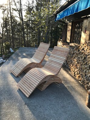 Two wooden lounge chairs in a sunlit outdoor area with a pile of firewood and a building with a blue awning in the background. - Cabin 'Kappacher' with Mountain View, Private Terrace and Wi-Fi (Bad Vigaun)