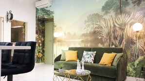 Ein Zimmer mit einem grünen Sofa, zwei Sesseln, einem Glastisch und einem Wandgemälde mit einer tropischen Szene.
