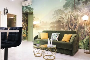Ein Zimmer mit einem grünen Sofa, zwei Sesseln, einem Glastisch und einem Wandgemälde mit einer tropischen Szene.