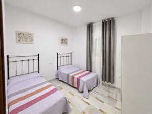 A bedroom with two beds, a window with curtains, a radiator, and a door. - Apartamento San ignacio de loyola, sol y ola I (Conil de la Frontera)