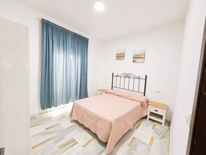 Un dormitorio con cama, mesita de noche, cortina azul y dos cuadros en la pared.