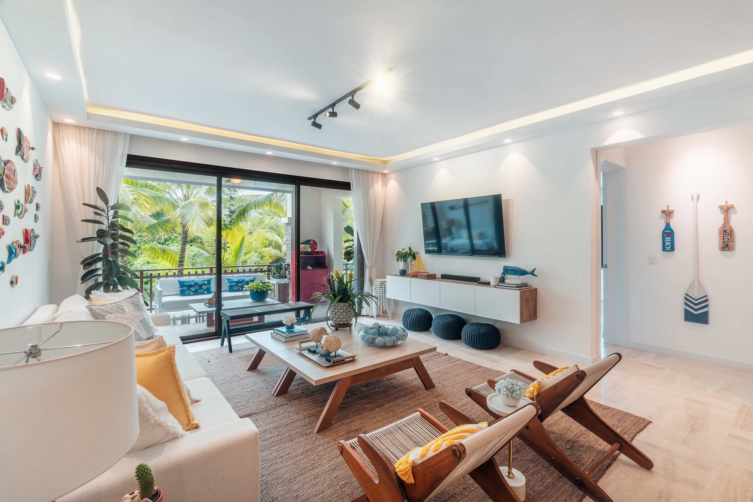 A modern living room with a sofa, coffee table, and lounge chairs. There's a TV mounted on the wall, a balcony with plants, and a large window.
