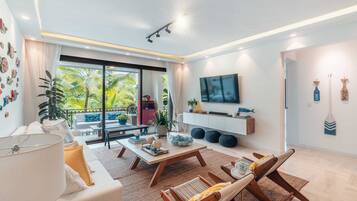 A modern living room with a sofa, coffee table, and lounge chairs. There's a TV mounted on the wall, a balcony with plants, and a large window.