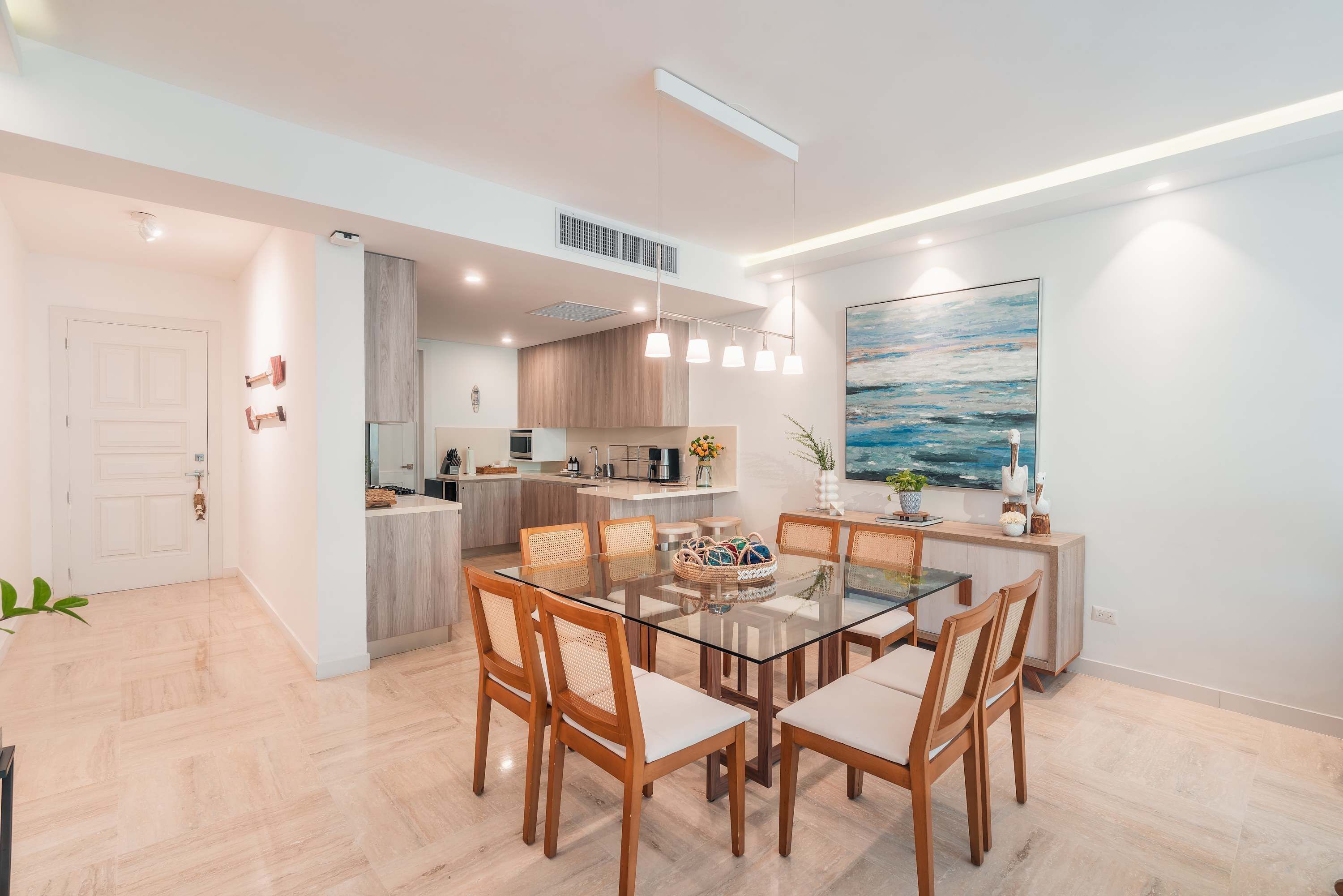 A modern kitchen with a central island, pendant lighting, and a dining area with a glass table and wooden chairs.