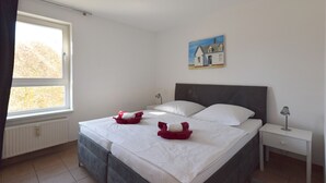 Un dormitorio con cama, dos almohadas, una mesita de noche, una lámpara, un cuadro y una ventana.