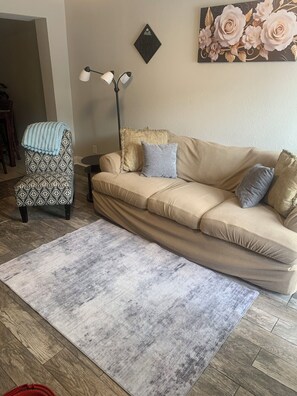 A living room with a patterned armchair, a beige sofa with cushions, a floor lamp, and a painting on the wall.