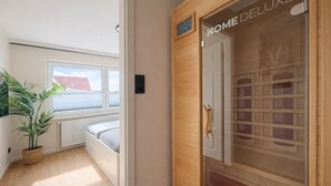 Una habitación con cama, una planta en maceta, ventana con vistas y la puerta de un sauna que dice 'HOME DELUXE'.