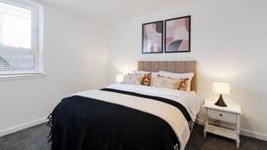 Een slaapkamer met een bed, een nachtkastje, een lamp, een raam met jaloezieën en twee abstracte schilderijen aan de muur.
