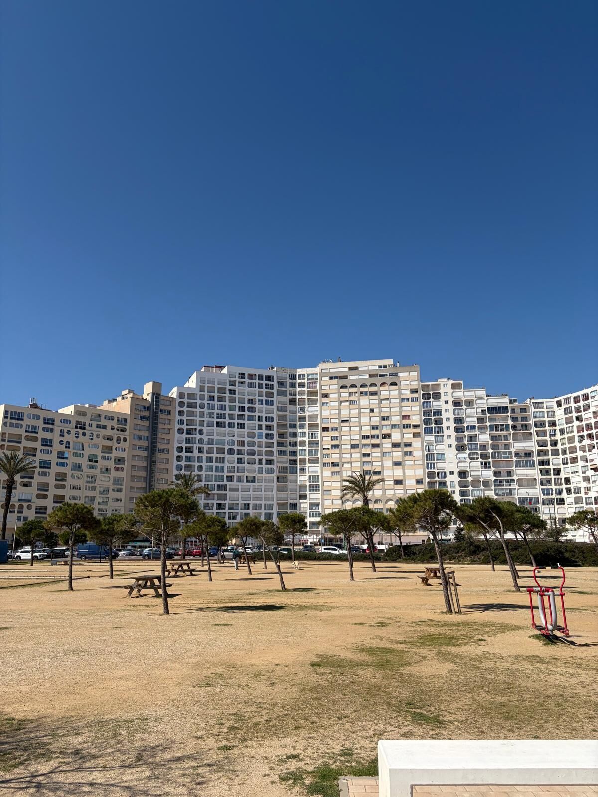 A high-rise hotel with multiple balconies, a sandy ground with sparse trees, and a clear blue sky.